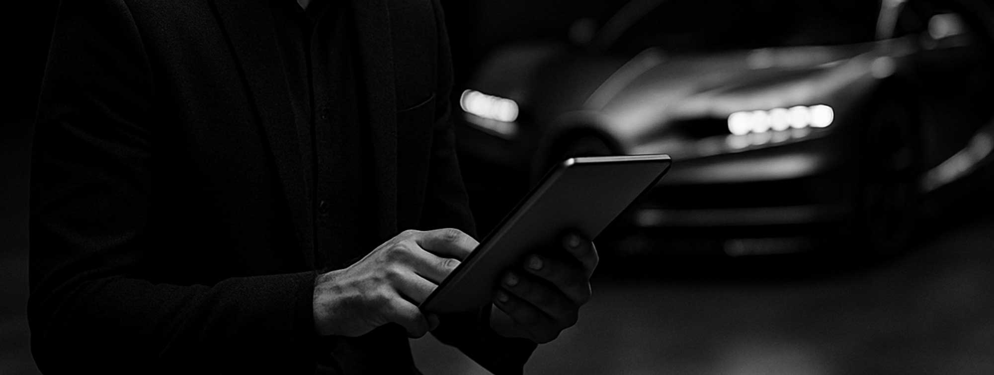 Black and white photo of a man reading the news on his tablet with a supercar in the background