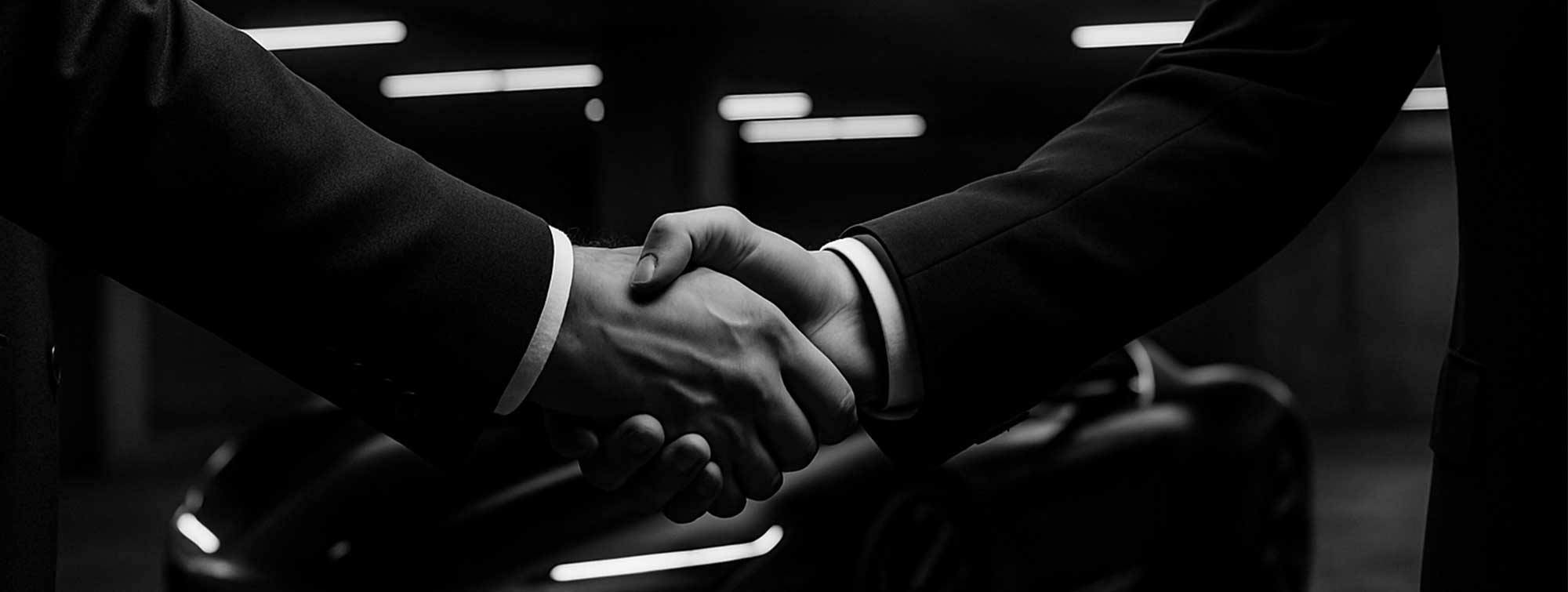 Black and white photo of two men in suits shaking hands with a supercar in the background in a chic garage lit by neon lights.