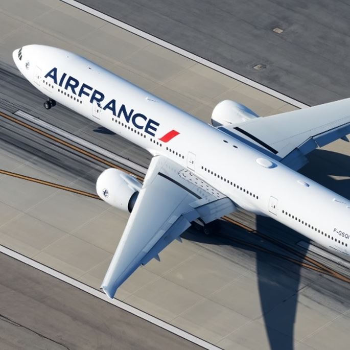 Homme en manteau élégant traversant un terminal d’aéroport moderne avec une valise cabine, avion visible sur le tarmac à travers la baie vitrée, en noir et blanc.