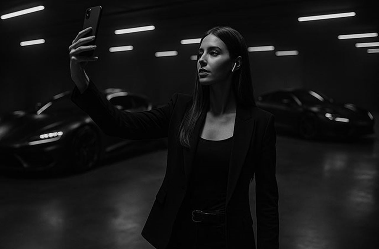 Black and white photograph of an influencer in a garage lit by neon lights, taking a selfie in front of a supercar.